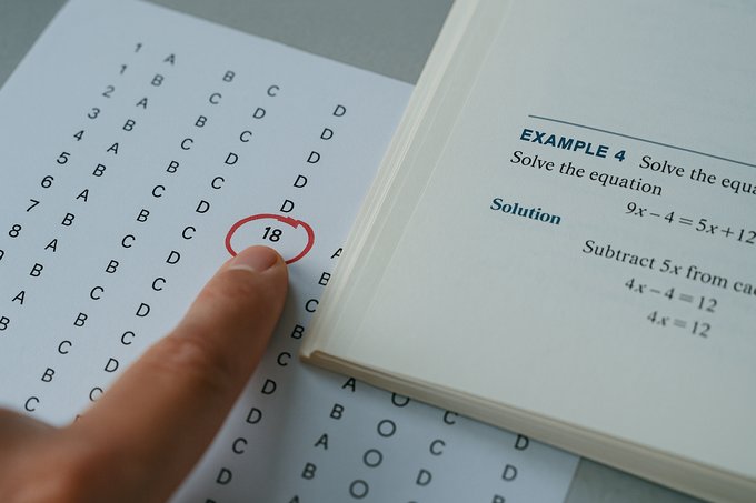 A fingertip touching a circled answer on an exam sheet next to an open textbook showing a similar worked example